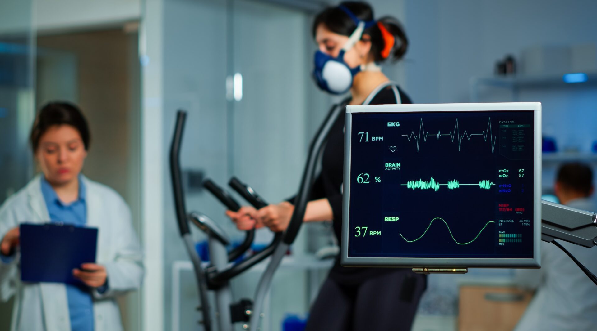Monitor showing EKG scan of athlete running on cross trainer and medical specialists supervise exercise in background, controlling physical activity, measuring heart rate in sports science laboratory